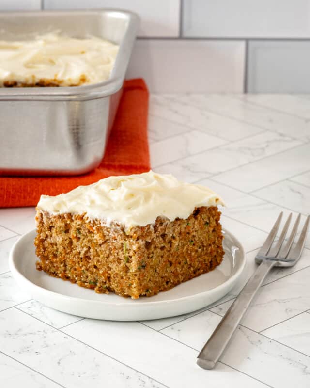 A zucchini carrot cake slice on a white plate with cream cheese frosting.
