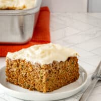 A zucchini carrot cake slice on a white plate with cream cheese frosting.