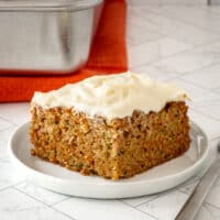 A zucchini carrot cake slice on a white plate with cream cheese frosting.