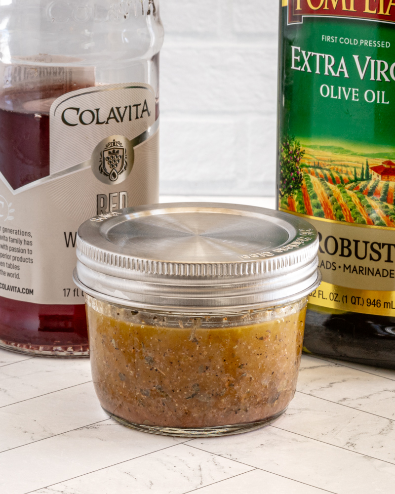 Freshly prepared seasoned meat mixture in a glass jar with olive oil and canned tomatoes in the background.