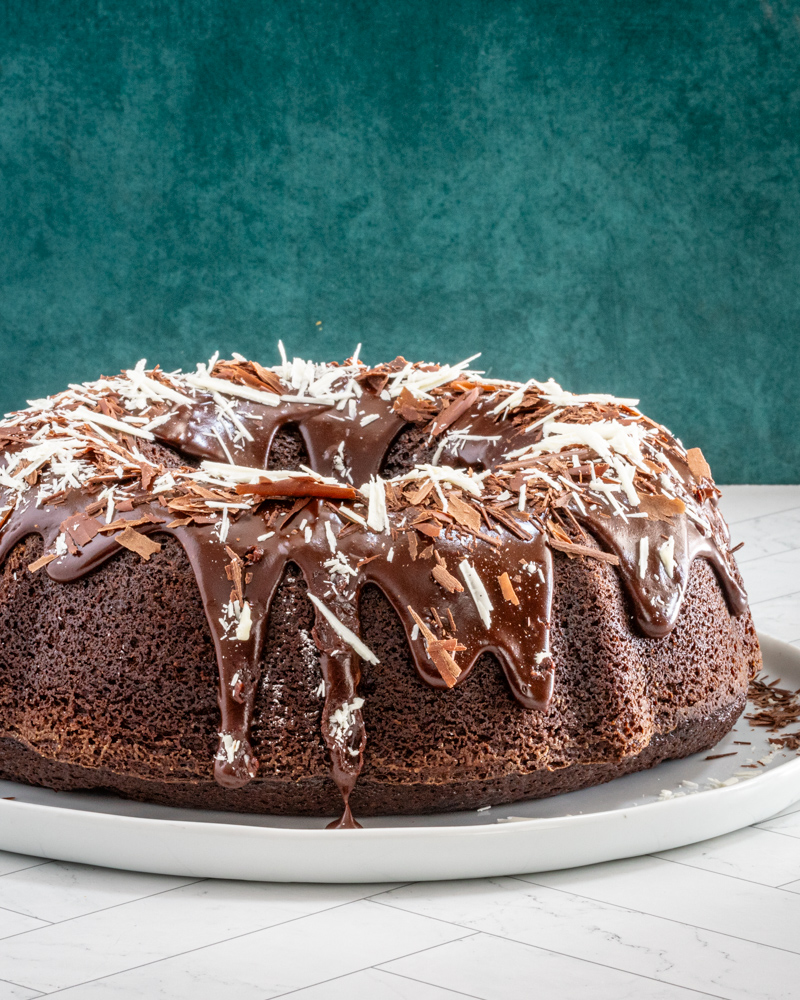 Moist chocolate bundt cake on a plate, decorated with a chocolate glaze and chocolate shards.