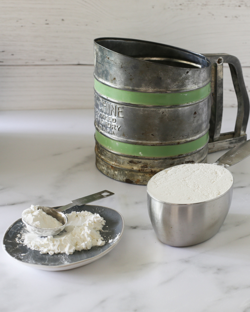 Flour in a sifter, measuring cup, and on a plate with measuring spoon.