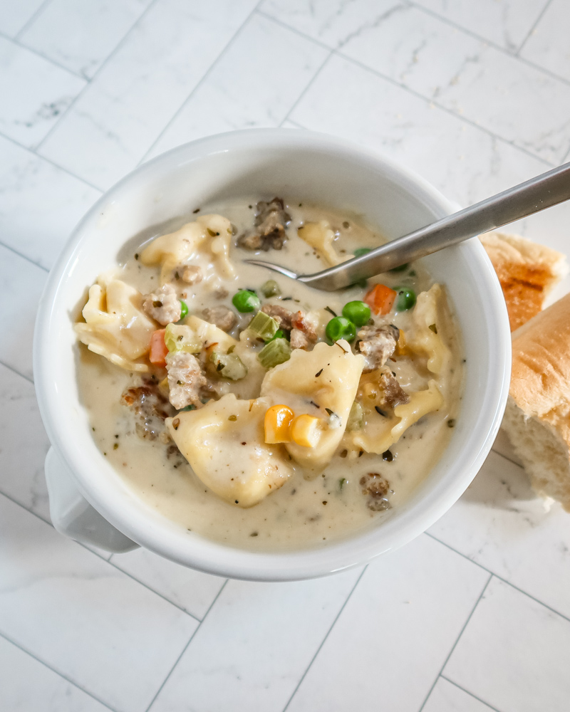 A bowl of sausage and tortellini soup with bread on the side.
