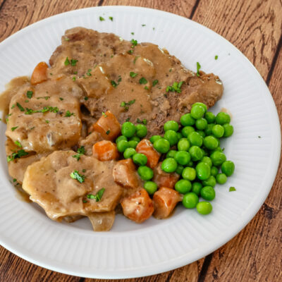 Cube steaks with gravy on a plate with peas, carrots, and sliced potatoes.