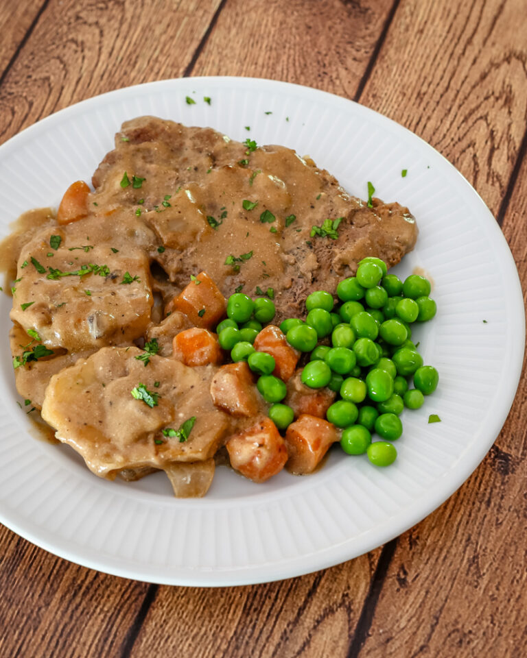 Cube steaks with gravy on a plate with peas, carrots, and sliced potatoes.