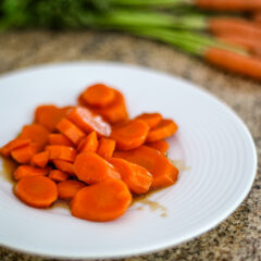 Glazed carrots on a white plate with a bunch of fresh carrots in the background.