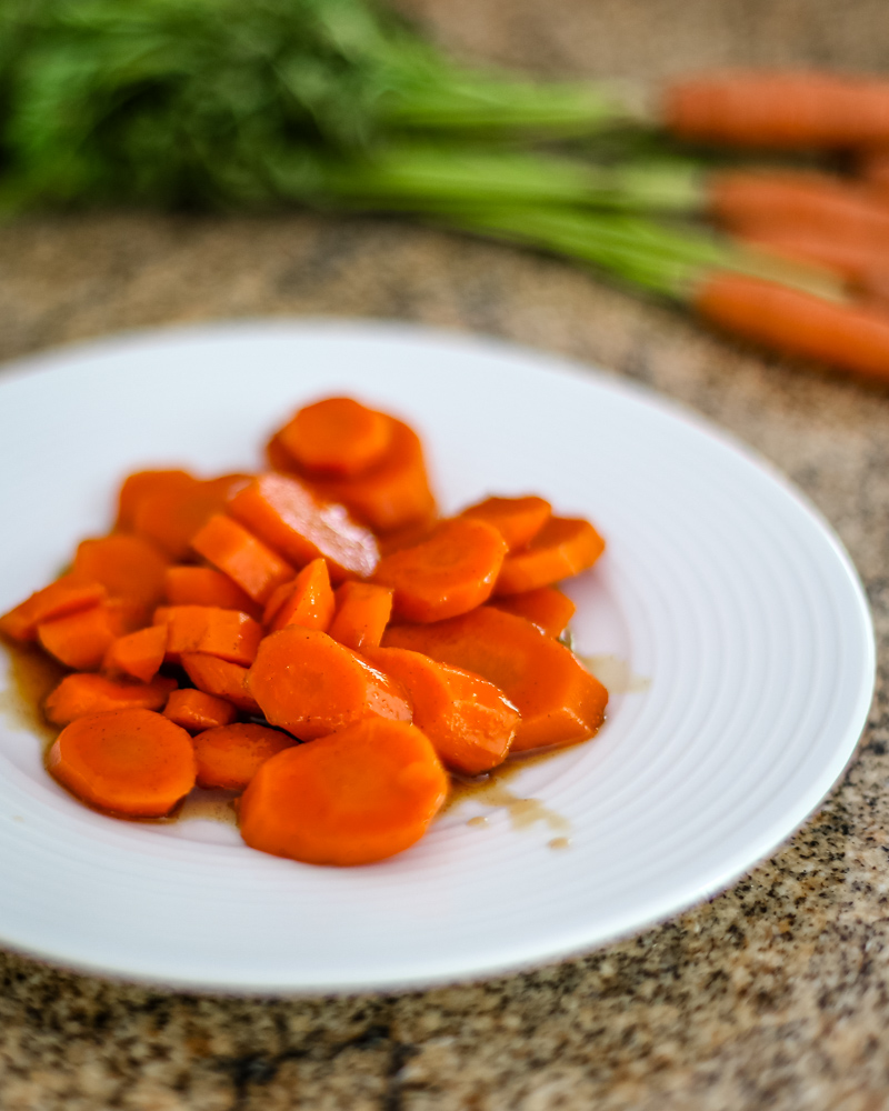 Glazed carrots on a white plate with a bunch of fresh carrots in the background.