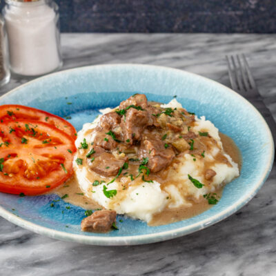 Beef tips on mashed potatoes with sliced tomatoes, on a blue plate.