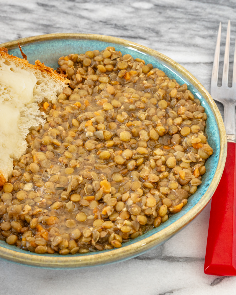 Bowl of cooked lentils with carrots and broth, with a piece of bread on the side.