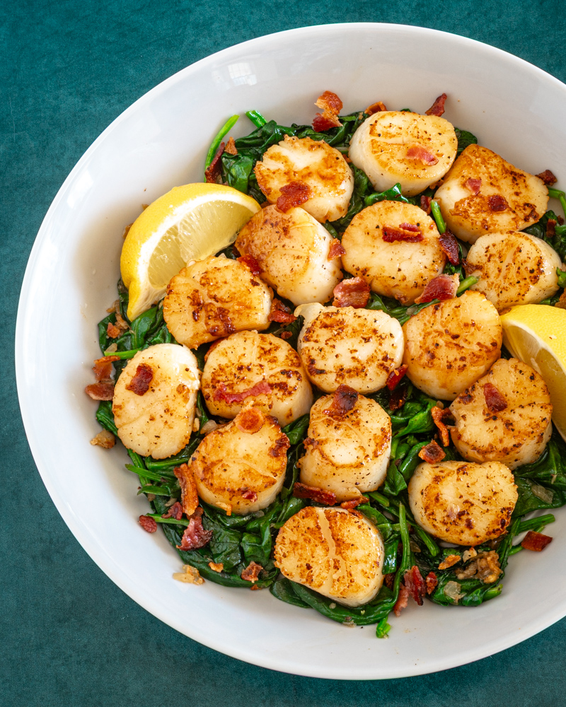 scallops on a bed of spinach with lemon wedges and bits of bacon