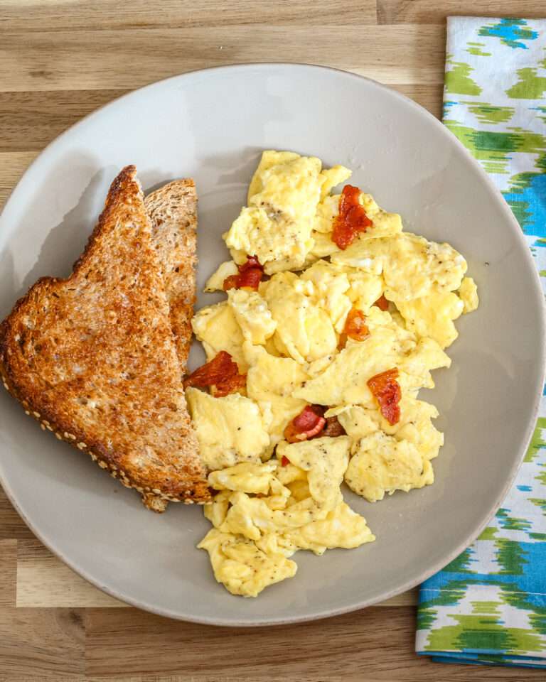 A plate of scrambled eggs with bacon and slices of toast on the side