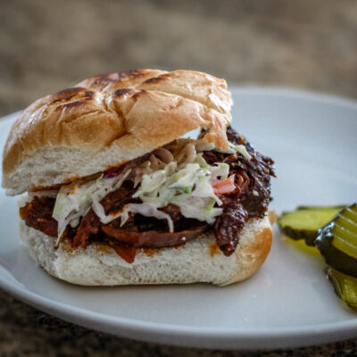 Slow-cooked shredded beef sandwich with coleslaw on a bun, garnished with pickles.