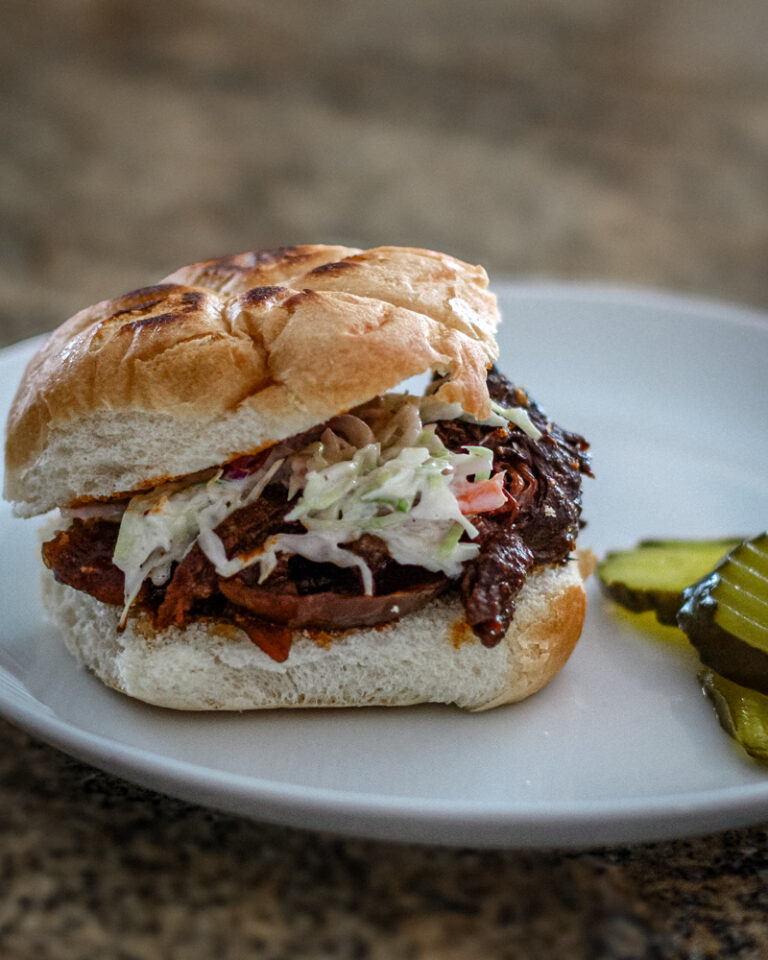 Slow-cooked shredded beef sandwich with coleslaw on a bun, garnished with pickles.