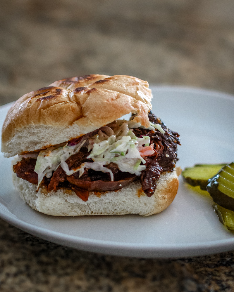 Slow-cooked shredded beef sandwich with coleslaw on a bun, garnished with pickles.