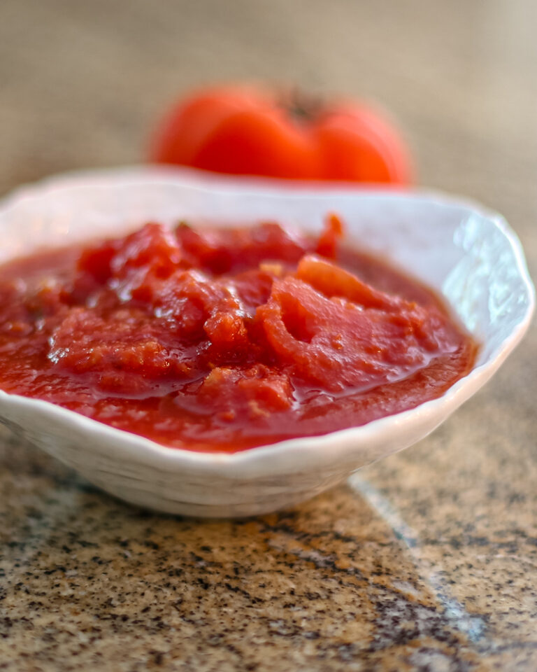 Homemade stewed tomatoes in a white serving bowl with a tomato in the background.