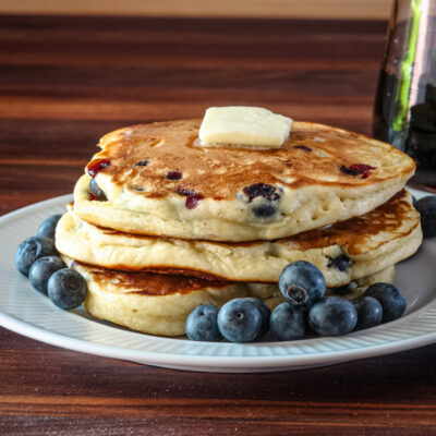 A stack of blueberry pancakes with some fresh blueberries, and a pat of butter, on a plate.