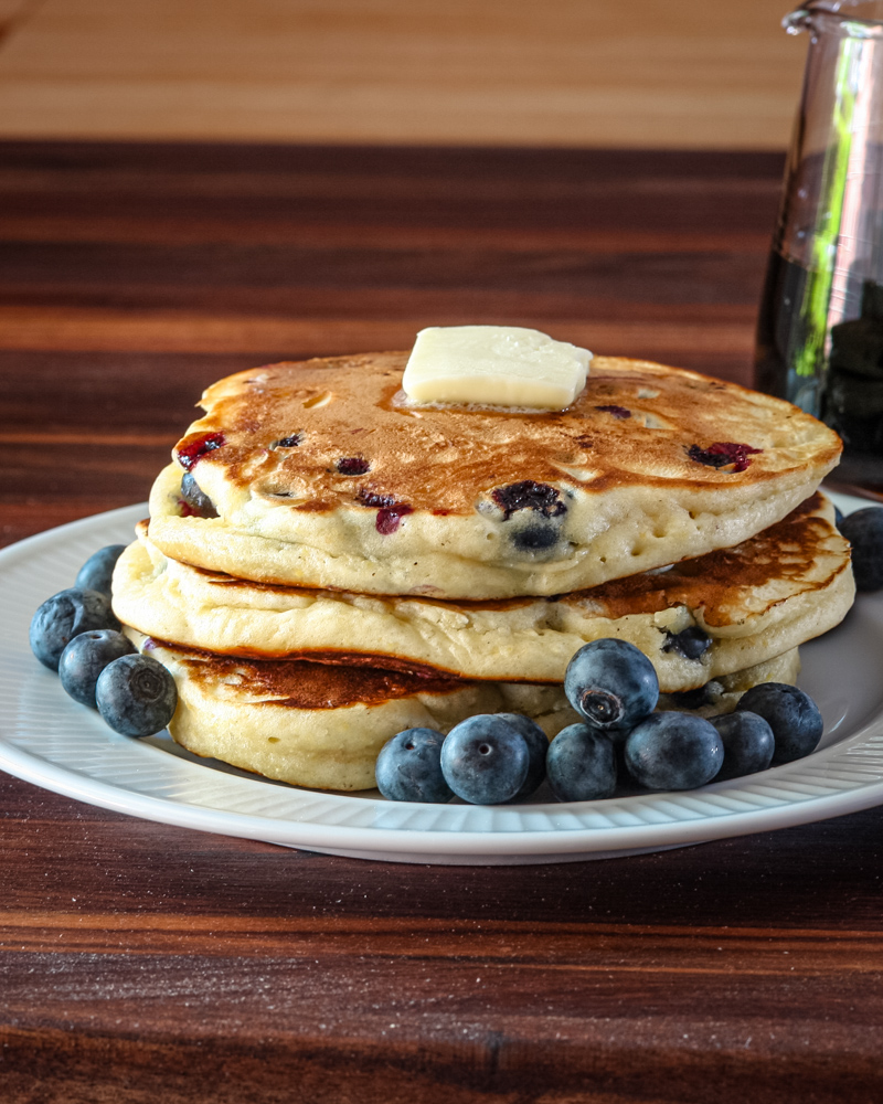 A stack of blueberry pancakes with some fresh blueberries, and a pat of butter, on a plate.