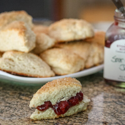 Scones with jam on the counter, with a plate of scones and jam jar in the background.