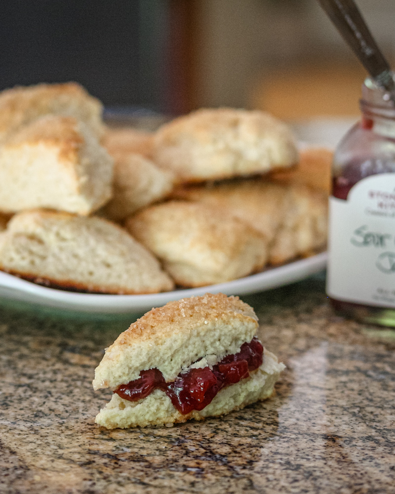 Scones with jam on the counter, with a plate of scones and jam jar in the background.
