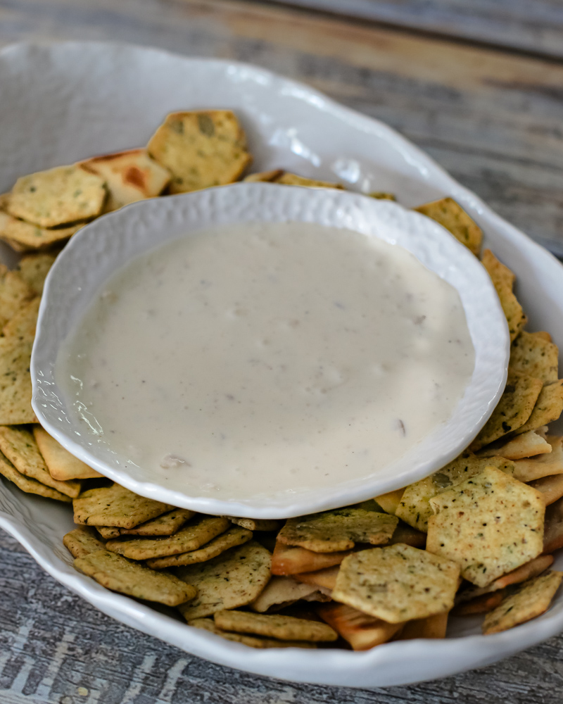 Creamy white clam dip in a white bowl surrounded by seasoned crackers on a rustic wooden surface.