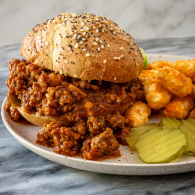 Sloppy Joe sandwich on a plate with some tater tots and pickles.