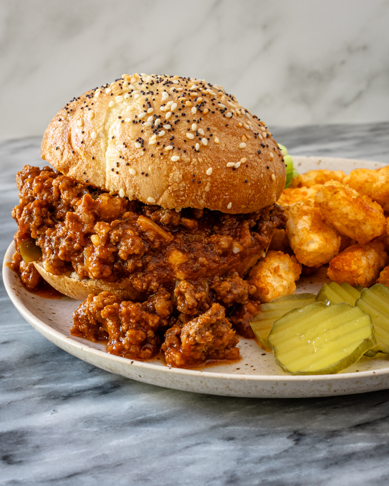 Sloppy Joe sandwich on a plate with some tater tots and pickles.