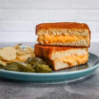 A grilled cheese sandwich from the air fryer, sliced and arranged on a plate with pickles and potato chips.
