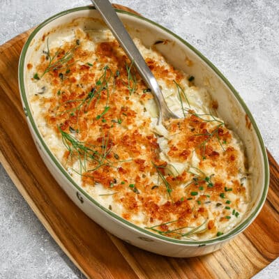 A creamy fennel gratin in an oval baking dish with a large spoon, on a cutting board and showing a crunchy fresh breadcrumb topping.