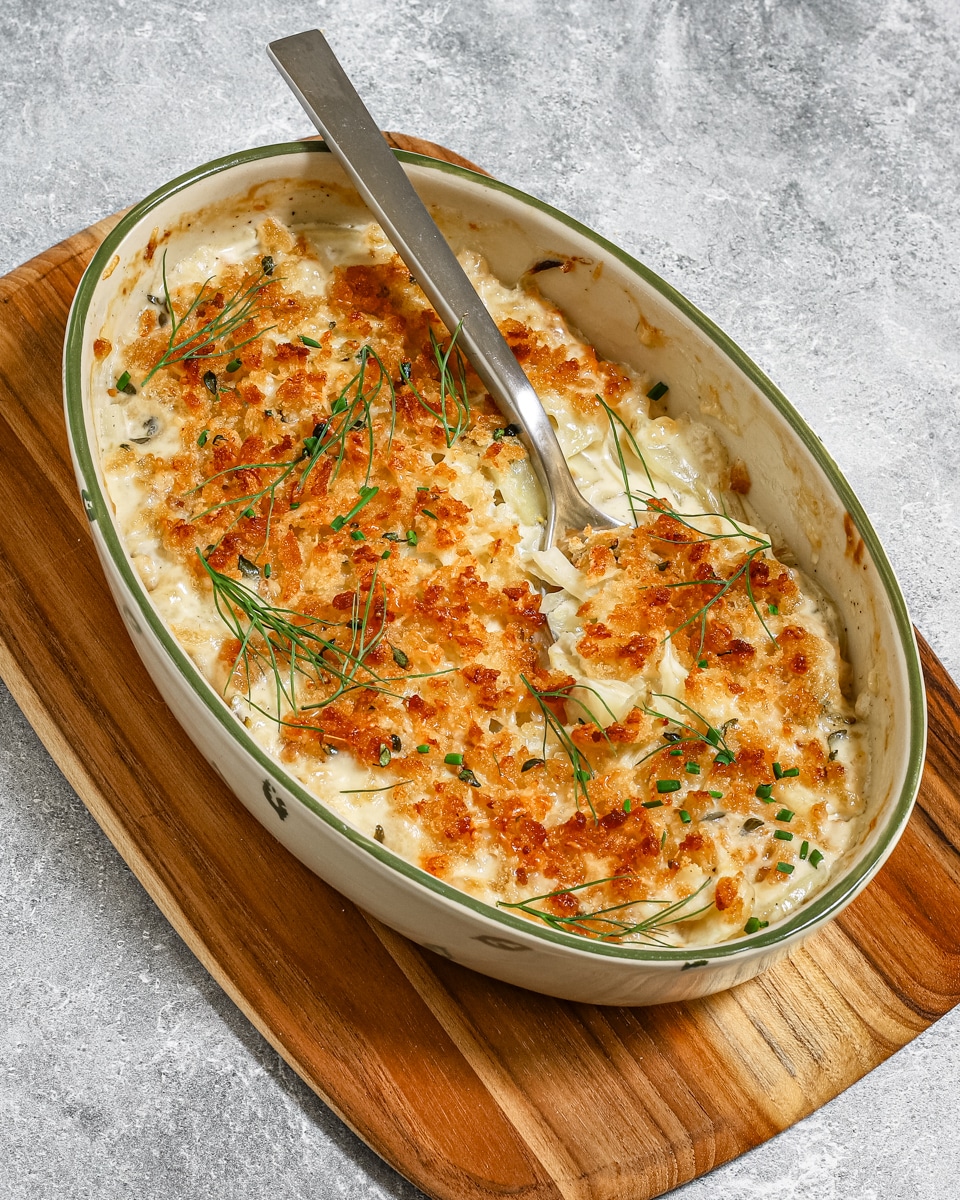 A creamy fennel gratin in an oval baking dish with a large spoon, on a cutting board and showing a crunchy fresh breadcrumb topping.