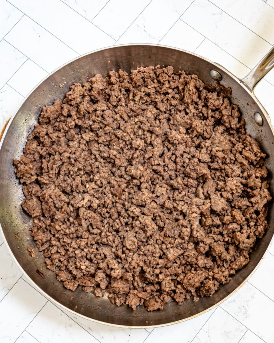 Browned ground beef in a large stainless steel skillet with a wooden spoon.
