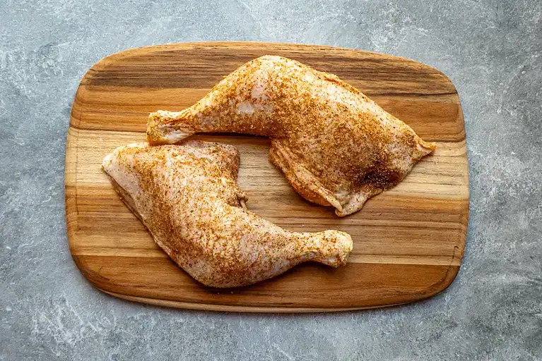 Chicken leg quarters on a small cutting board being seasoned and ready to put in the air fryer.