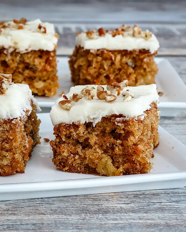 Moist carrot cake squares with cream cheese frosting and chopped nuts on top.