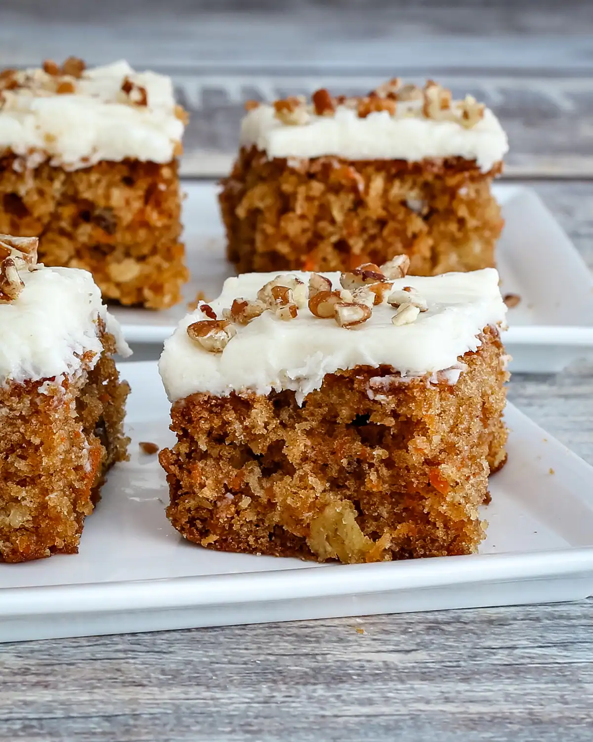 Moist carrot cake squares with cream cheese frosting and chopped nuts on top.