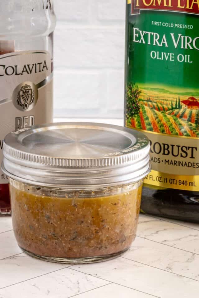 Freshly prepared seasoned meat mixture in a glass jar with olive oil and canned tomatoes in the background.