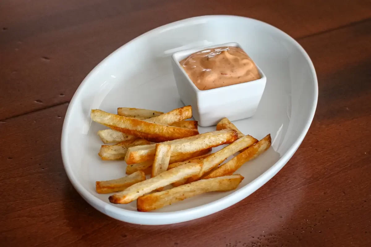Crispy French fries with dipping sauce in a white dish on wooden table.