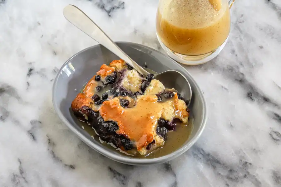 A dessert dish with blueberry bread pudding and vanilla sauce on the side.