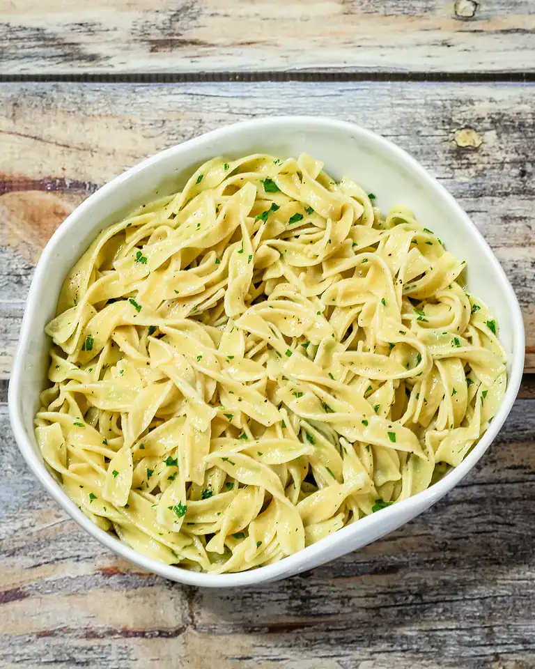 Creamy garlic pasta with parsley in a white bowl on rustic wooden surface.