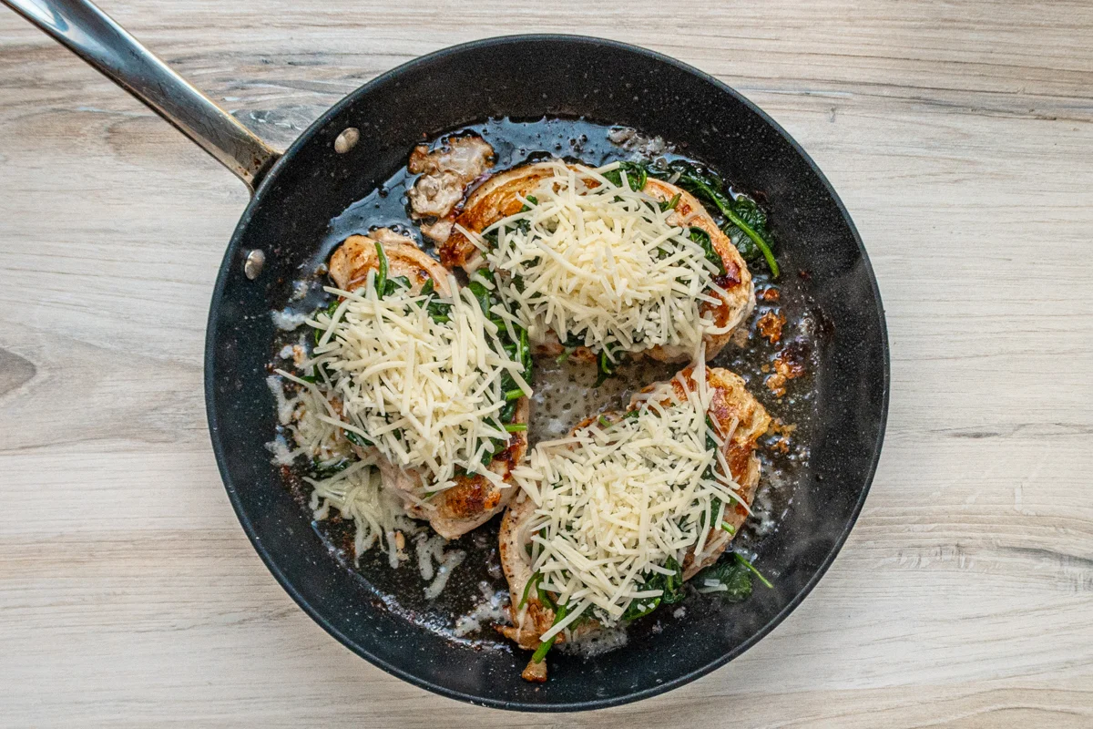 Chicken breasts in the skillet topped with wilted spinach and shredded Gruyère cheese.