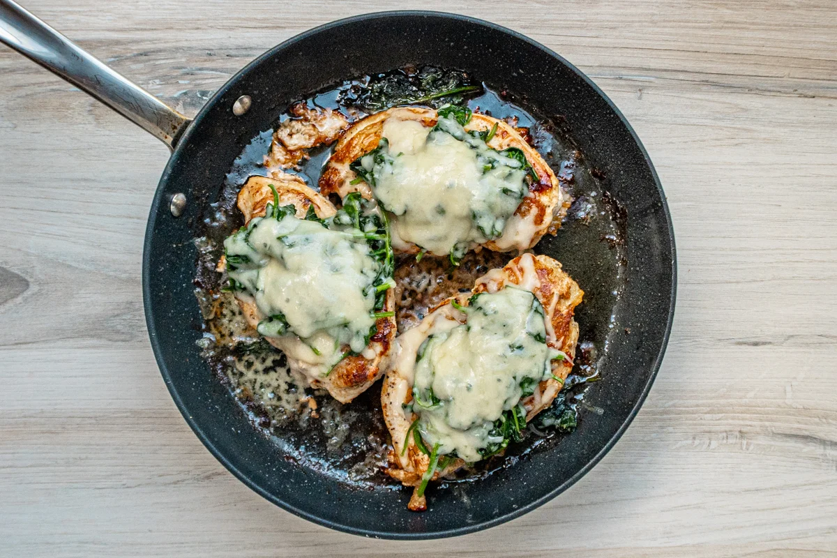 Chicken breasts in the skillet topped with wilted spinach and melted Gruyère cheese.