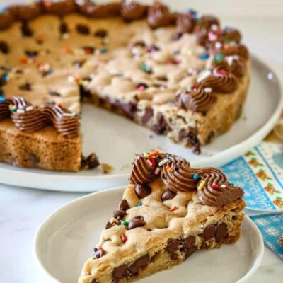 A slice of chocolate chip cookie cake with piped chocolate frosting and rainbow sprinkles.