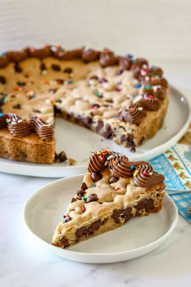 A slice of chocolate chip cookie cake with piped chocolate frosting and rainbow sprinkles.
