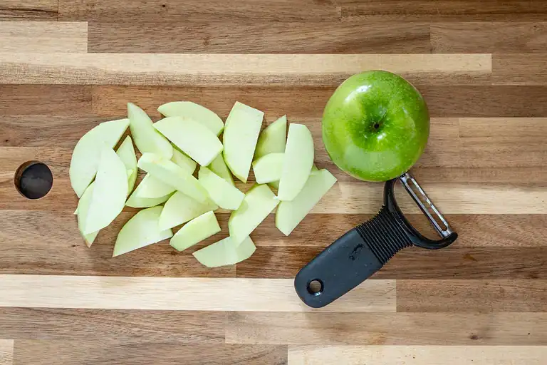 Sliced green apple on wooden cutting board with apple peeler.