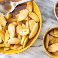 Baked apple slices in caramel sauce in yellow bowls on marble surface.