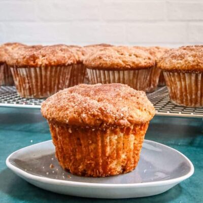 A cinnamon muffin on a small plate with the rack of muffins behind it.