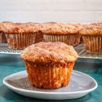 A cinnamon muffin on a small plate with the rack of muffins behind it.