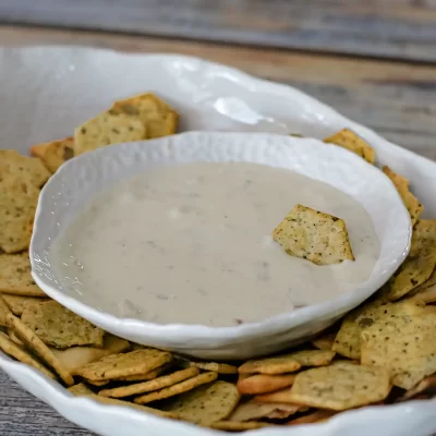 Creamy white dip in a white bowl surrounded by seasoned crackers on a rustic wooden surface.