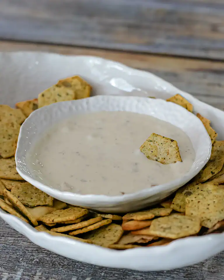 Creamy white dip in a white bowl surrounded by seasoned crackers on a rustic wooden surface.