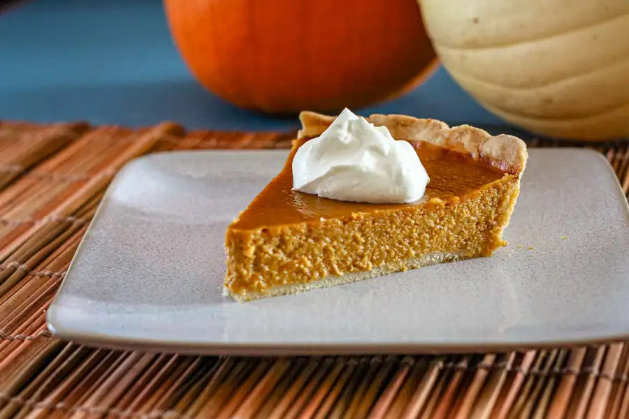 A slice of classic pumpkin pie on a plate with a dollop of whipped cream and small pumpkins in the background.