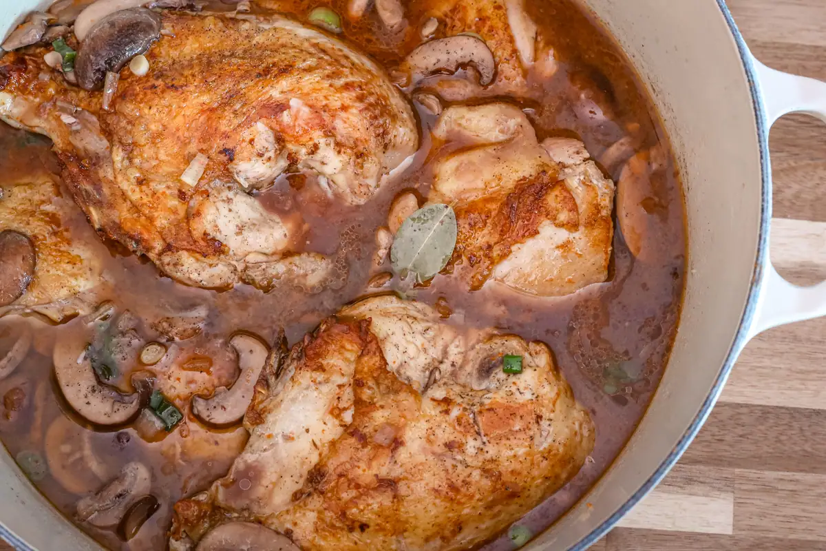 Coq au Vin chicken dish in a Dutch oven with mushrooms and bay leaf.