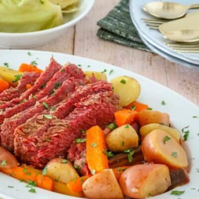 corned beef on a platter with potatoes, carrots, and cabbage on a plate in the background
